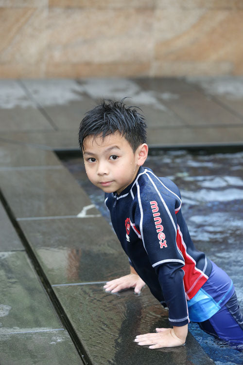 一個頭髮濕漉漉的小男孩,穿著藏青色和紅色相間的游泳襯衫,正從泳池中爬到濕漉漉的石面上,看向攝影機的方向。.